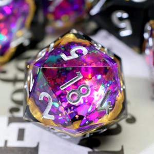 May include: A close-up of a multi-faceted, clear resin die with silver numbering. The die is filled with purple, pink, and gold glitter. The numbers 2, 4, 5, and 18 are visible. Other dice are visible in the background.