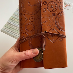 May include: A brown leather journal with clock designs embossed on the cover. The journal is closed with a leather cord and a clock charm. The cover features Roman numerals and clock hands. A small piece of paper with text is visible.