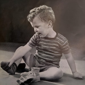 Puede incluir: Fotografía en blanco y negro de un niño pequeño sentado en el suelo. El niño lleva una camiseta de manga corta a rayas, pantalones cortos y calcetines. Un zapato está en el suelo cerca del niño.