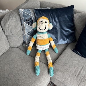 Peut inclure: Une peluche singe crochetée à la main présentant un design coloré en jaune moutarde, crème et bleu sarcelle. Le singe a un visage souriant avec des yeux et une bouche brodés en noir, de longs membres et un détail blanc rappelant une couche.