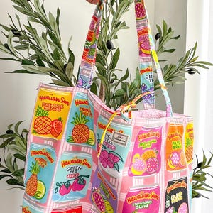 May include: A fabric tote bag featuring a repeating grid pattern of colorful Hawaiian Sun drink cans in various flavors like Pineapple Orange, Strawberry Guava, and Luau Punch. The bag has two matching fabric handles and a light pink checkered background.
