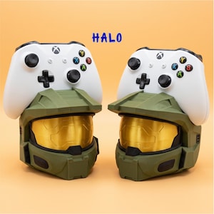 May include: Two white game controllers resting on top of two olive green helmet stands with gold visors. The word HALO is printed in blue text above the controllers. The helmet stands feature a visor design and a matte finish on the green surface.