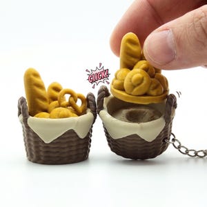 Op de afbeelding: Twee miniatuur sleutelhangers in de vorm van bruine manden gevuld met brood, pretzels en broodjes. Eén sleutelhanger heeft een deksel dat wordt opgetild en de inhoud onthult. Het woord "CLICK!" staat in een tekstballon. Witte achtergrond.