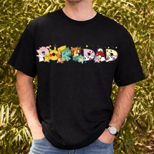 May include: A black short-sleeved t-shirt featuring the text POKEDAD in large, colorful letters. Each letter is filled with various cartoon characters and patterns, including Pikachu, Charmander, Squirtle, and Jigglypuff, accented by small stars.