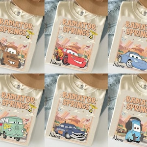 May include: Cream-colored t-shirts featuring various cartoon car characters from the movie "Cars." Each shirt displays "Radiator Springs" text and a space for a name. The designs include Mater, Lightning McQueen, Sally, and other vehicles.