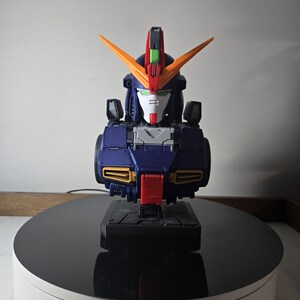 May include: A detailed model of a robot bust, featuring a color scheme of navy blue, orange, red, and white. The robot has a distinctive head design with V-shaped orange accents and is mounted on a black base.