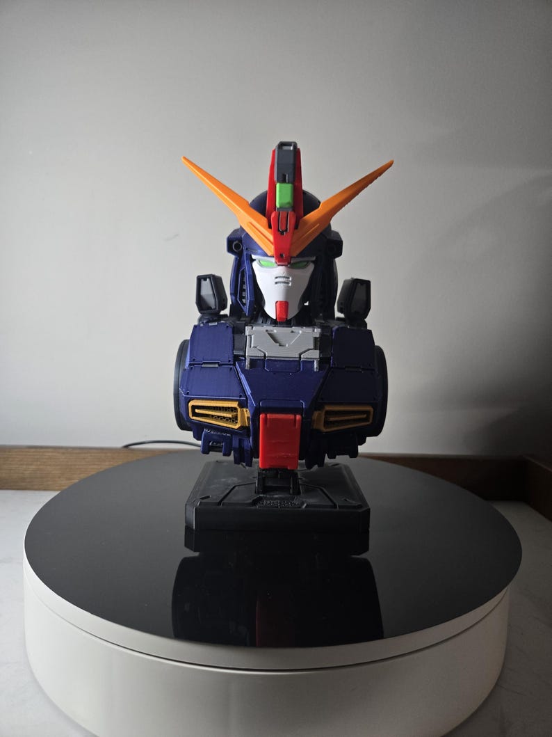 May include: A detailed model of a robot bust, featuring a color scheme of navy blue, orange, red, and white. The robot has a distinctive head design with V-shaped orange accents and is mounted on a black base.