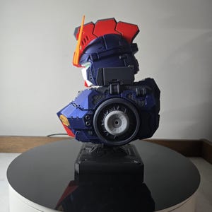 May include: A detailed model of a robot's head and upper torso, featuring a color scheme of blue, red, and white. The model has intricate mechanical details, including a circular component on the side. It is displayed on a black rotating platform.