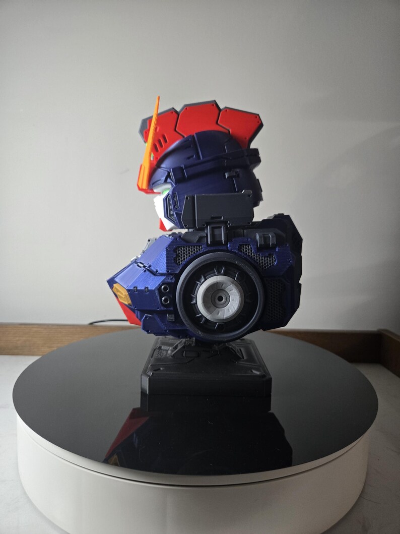 May include: A detailed model of a robot's head and upper torso, featuring a color scheme of blue, red, and white. The model has intricate mechanical details, including a circular component on the side. It is displayed on a black rotating platform.
