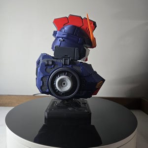 May include: A detailed model of a robot's head and upper torso, featuring a color scheme of navy blue, red, orange, and white. The model is mounted on a black base and displayed on a rotating platform.