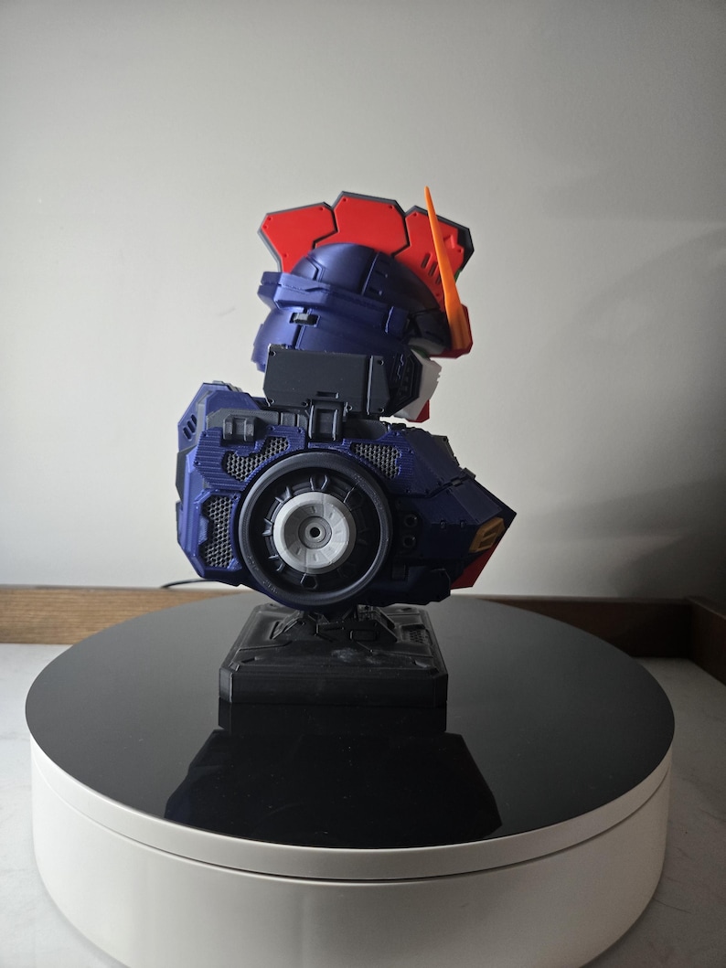 May include: A detailed model of a robot's head and upper torso, featuring a color scheme of navy blue, red, orange, and white. The model is mounted on a black base and displayed on a rotating platform.