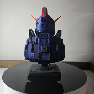 May include: A dark blue robot model with orange and red accents. The model is composed of geometric shapes and is mounted on a black base. The robot is displayed on a rotating platform with a white base and a black top.