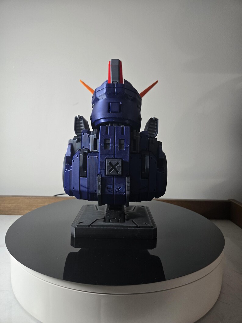 May include: A dark blue robot model with orange and red accents. The model is composed of geometric shapes and is mounted on a black base. The robot is displayed on a rotating platform with a white base and a black top.