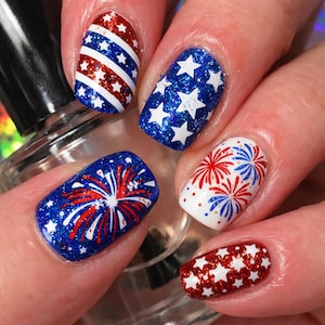 May include: Close-up of a hand with a manicure featuring patriotic nail art. Designs include red, white, and blue glitter, stars, stripes, and fireworks. The nails are painted in various shapes and sizes.