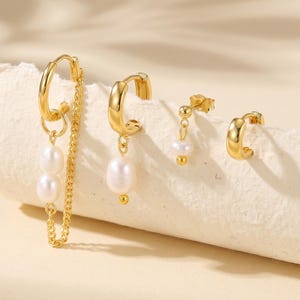 May include: A collection of gold-colored earrings featuring various designs. One pair has a chain with two dangling pearls, another has a single pearl pendant, a third has a small pearl, and the last is a small hoop. The earrings are displayed on a white surface.