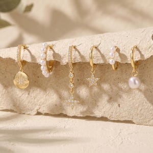 May include: A selection of gold-coloured hoop earrings with different charms. The charms include a starburst, pearls, a dangling star, and a single pearl. Some hoops are encrusted with small, clear stones.