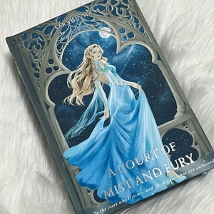 Puede incluir: Libro de tapa dura titulado "A Court of Mist and Fury" de Sarah J. Maas. La portada presenta a una mujer con un vestido azul fluido, sobre un fondo de cielo estrellado. El título del libro está en letras plateadas.