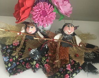 May include: Two handmade dolls dressed in colorful fabric with floral patterns, holding a stick and wearing a hat. The dolls are surrounded by pink, red, and purple flowers.