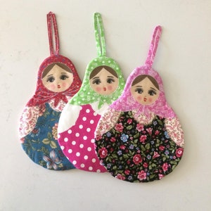 May include: Three handmade fabric ornaments shaped like Russian nesting dolls. The dolls are decorated with different floral patterns and have painted faces.