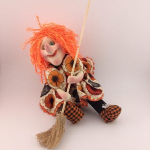 Può includere: Bambola di strega fatta a mano con capelli di filo arancione, un viso sorridente e un vestito a fantasia floreale. La bambola tiene una scopa e indossa scarpe a scacchi arancioni e nere, perfetta per Halloween.