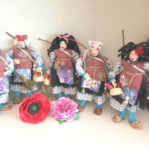Può includere: Una collezione di bambole di arte popolare fatte a mano, ognuna vestita con un grembiule patchwork unico, un maglione rosso e un copricapo fantasia. Alcune bambole portano cesti o scope, con un papavero rosso e un fiore rosa in primo piano.
