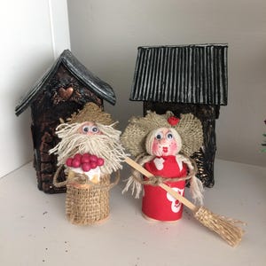 May include: Two handmade figurines of a man and woman dressed in rustic clothing. The man is holding a basket of red berries and the woman is holding a broom. They are standing in front of two small houses with brown and black roofs.