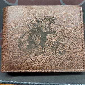 May include: Brown leather wallet featuring a black Godzilla graphic. The wallet has a textured surface and a bi-fold design. The Godzilla image is centered on the front of the wallet.