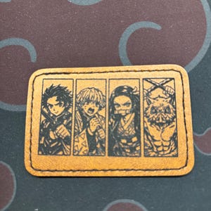 May include: A rectangular, tan leather patch featuring four black illustrations of anime characters. Each character is in a separate frame. The patch has a stitched border and is likely a decorative item or accessory.