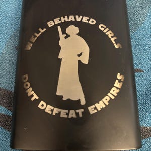 Puede incluir: Una petaca de metal negro mate con una silueta grabada a láser en tono plateado de una figura que sostiene un bláster. El texto "WELL BEHAVED GIRLS DONT DEFEAT EMPIRES" está grabado en un patrón circular alrededor de la figura central frontal.