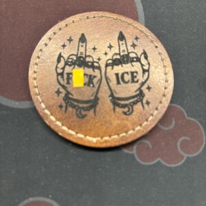 Puede incluir: Un parche redondo de cuero marrón con borde cosido. Presenta dos manos con los dedos medios levantados, con las palabras "F*CK" e "ICE". El diseño incluye estrellas, lunas y anillos.
