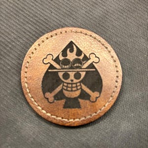 May include: A round, brown leather patch featuring a black and white skull and crossbones design with a flaming ace of spades. The patch is stitched around the edge and set against a dark gray background.