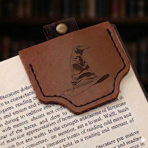 May include: Brown leather bookmark featuring a detailed engraving of the Sorting Hat from the Harry Potter series. The bookmark has a stitched border and a small strap with a metal rivet. It is placed on the pages of a book.