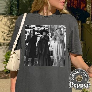 May include: Dark gray t-shirt featuring a black and white image of three figures. The image is a still from a movie. The shirt has the words "COMFORT COLORS" and "Pepper" printed on the bottom right.
