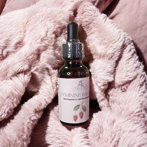 Könnte beinhalten: Eine dunkel bernsteinfarbene Glastropfflasche mit einem weißen Etikett, das den Text Vera & Vine, Feminine Balance und Red Raspberry Leaf Botanical Blend sowie eine Abbildung von zwei roten Himbeeren an einem Stiel mit grünen Blättern zeigt.