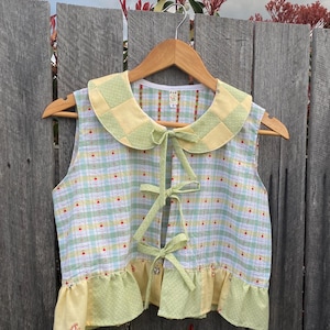 May include: A sleeveless vest featuring a light green, yellow, and white plaid pattern with small red accents. The garment has a large rounded collar, three front tie closures made of green fabric, and a ruffled hem in yellow and green.