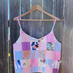 Könnte beinhalten: Ein ärmelloses Camisole-Top mit einem Patchwork-Design aus verschiedenen Stoffquadraten in Rosa-, Hellblau- und Violetttönen. Das Muster umfasst Vichy-Karos, einfarbige Flächen und Blumenprints, ergänzt durch dünne Schulterträger.