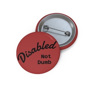 Puede incluir: Un botón rojo con las palabras "Disabled Not Dumb" en escritura negra. El botón tiene un respaldo de alfiler de metal y un borde plateado. El botón tiene forma redonda.