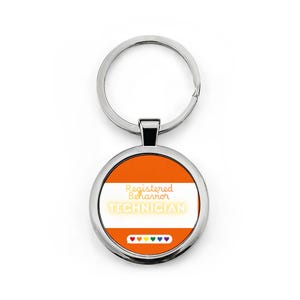 Op de afbeelding: Een ronde zilveren sleutelhanger met een oranje en wit gestreepte achtergrond. De tekst luidt Registered Behavior TECHNICIAN in witte en gele letters. Onder de tekst staat een wit tekstballonpictogram met zes kleine gekleurde hartjes.