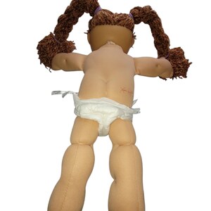 Puede incluir: Una mu&ntilde;eca vintage con coletas de hilo marr&oacute;n sujetas con bandas moradas. La mu&ntilde;eca tiene un tono de piel claro y lleva un pa&ntilde;al. Los brazos de la mu&ntilde;eca est&aacute;n extendidos y la espalda es visible.