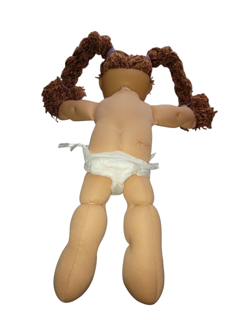 Puede incluir: Una mu&ntilde;eca vintage con coletas de hilo marr&oacute;n sujetas con bandas moradas. La mu&ntilde;eca tiene un tono de piel claro y lleva un pa&ntilde;al. Los brazos de la mu&ntilde;eca est&aacute;n extendidos y la espalda es visible.