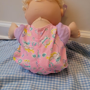 May include: A doll with blonde curly hair and a light skin tone, wearing a pink short-sleeved shirt with a repeating pattern of children playing with balls and jump ropes, featuring lavender sleeves and a small purple bow in the hair.
