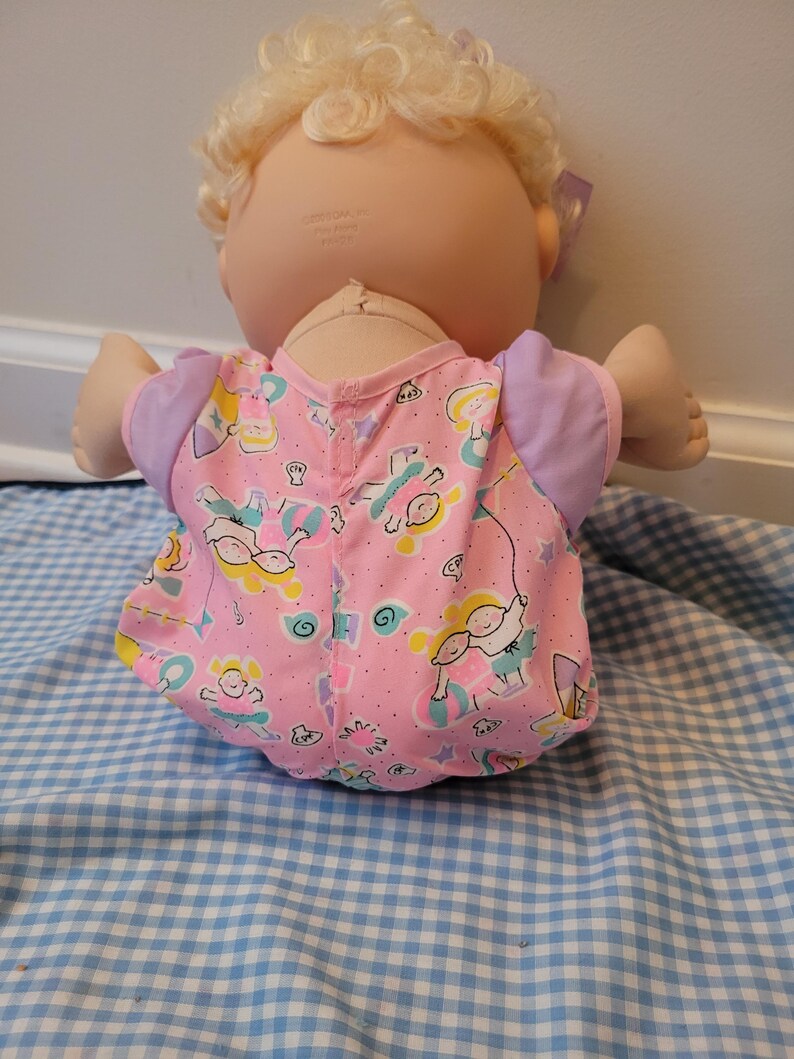 May include: A doll with blonde curly hair and a light skin tone, wearing a pink short-sleeved shirt with a repeating pattern of children playing with balls and jump ropes, featuring lavender sleeves and a small purple bow in the hair.