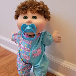 May include: A Cabbage Patch Kids doll with curly brown hair, green eyes, and a light blue pacifier. The doll wears a one-piece outfit featuring a solid light blue side and a patterned side with pink balloons, yellow stars, and the text CPK.