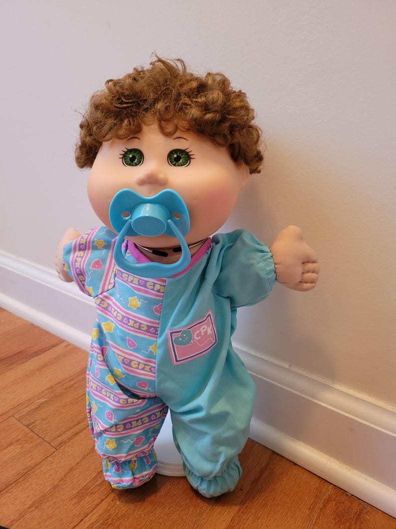 May include: A Cabbage Patch Kids doll with curly brown hair, green eyes, and a light blue pacifier. The doll wears a one-piece outfit featuring a solid light blue side and a patterned side with pink balloons, yellow stars, and the text CPK.