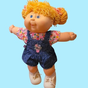 May include: A vintage Cabbage Patch Kid doll with blonde, curly hair and blue eyes. The doll wears a floral print shirt under denim overalls with a flower pot graphic. White sneakers complete the outfit.