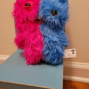 May include: A plush toy with a split design, half hot pink and half blue, with large, round eyes. The furry creature is perched on a light blue wooden chair, creating a whimsical and colorful display. The toy has a tag attached.