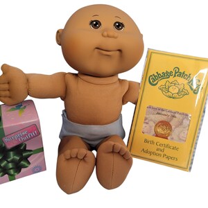 May include: A bald doll with brown skin and brown eyes wearing a light blue diaper, positioned next to a pink box labeled Surprise Outfit with a green bow and a yellow envelope labeled Cabbage Patch Kids Birth Certificate and Adoption Papers.