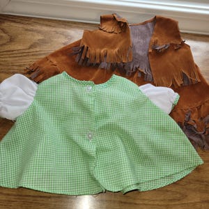 Op de afbeelding: Een groen-wit geruite cape met witte mouwen en een bruin su&egrave;de vest met franjes. De cape heeft een knoopsluiting. Het vest is bruin met een grijze voering en franjes langs de onderkant en zijkanten.