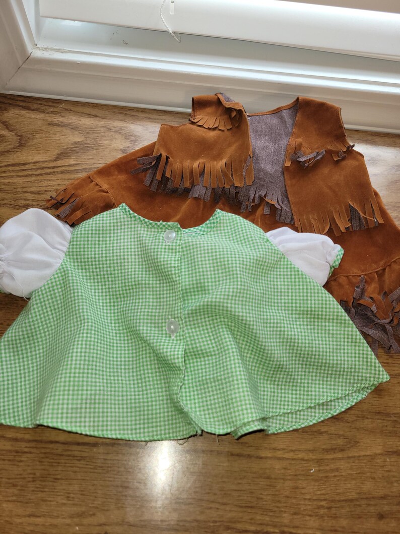 Op de afbeelding: Een groen-wit geruite cape met witte mouwen en een bruin su&egrave;de vest met franjes. De cape heeft een knoopsluiting. Het vest is bruin met een grijze voering en franjes langs de onderkant en zijkanten.