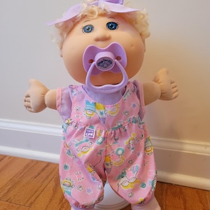 May include: A Cabbage Patch Kids doll with blonde curly hair, blue eyes, and a light purple bow. The doll wears a light purple shirt under pink overalls featuring a colourful pattern of small figures and stars, with a matching light purple pacifier.